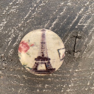 May include: A round, white, glass knob with a printed image of the Eiffel Tower and a pink flower. The knob has a glossy finish.