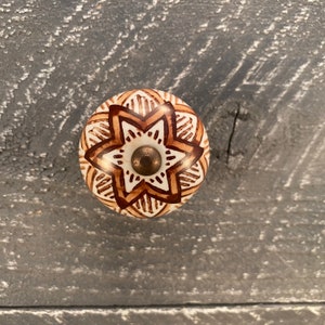 Può includere: Un pomello in ceramica bianca con un design a mandala marrone. Il pomello ha una vite in metallo dorato al centro.