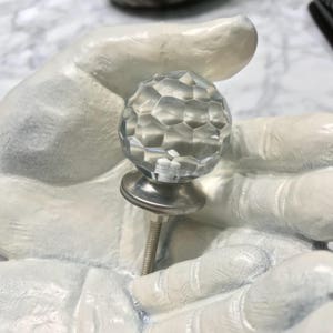 Puede incluir: Un pomo de armario de cristal transparente con un diseño facetado y una base de metal plateado y un tornillo.