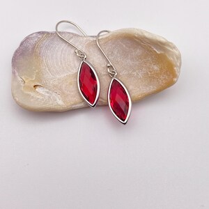 May include: A pair of silver-toned drop earrings with marquise-shaped, faceted, red gemstones. The earrings are displayed on a light-colored, textured seashell against a white background. The gemstones have a bright, reflective quality.