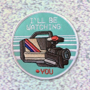 May include: A teal blue embroidered patch with a white border. The patch features a cartoon image of a vintage video camera with the text "I'll be watching - you".