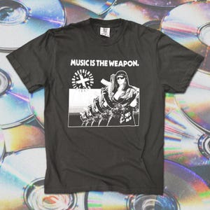 May include: Black t-shirt with the text "MUSIC IS THE WEAPON." and a graphic of a person holding a gun. The graphic is in black and white. The shirt is a crew neck.