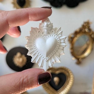 Peut inclure: Un pendentif blanc en forme de cœur avec un motif de couronne et d'éclat de soleil. Le pendentif est tenu devant un arrière-plan flou avec d'autres objets décoratifs, dont un papillon noir et des cœurs dorés.