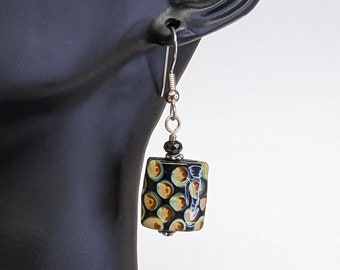 May include: A pair of dangle earrings with a black and brown rectangular ceramic bead with a white and green pattern. The bead is attached to a silver hook.
