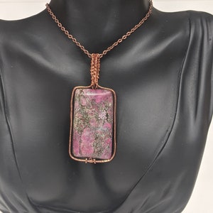 May include: A copper wire wrapped pendant necklace with a pink and green stone. The stone is rectangular and has a marbled pattern.