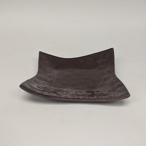 Puede incluir: Un plato pequeño, cuadrado, de cerámica marrón con textura martillada. El plato tiene bordes ligeramente redondeados y un borde ligeramente elevado.