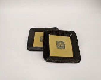 May include: Two square ceramic plates with black rims and a light yellow center. Each plate has a raised design of a stylized face in the center.