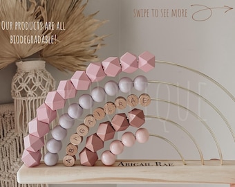 May include: A wooden abacus with pink geometric beads and wooden letter beads spelling "ABIGAIL RAE". The abacus has gold-colored metal arches and is displayed on a light-colored wooden base. The text "OUR PRODUCTS ARE ALL BIODEGRADABLE!" is visible.
