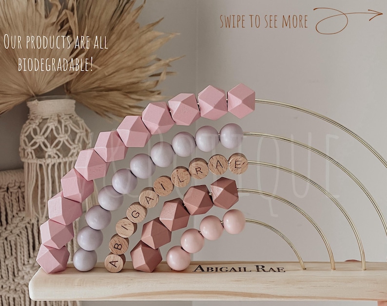 May include: A wooden abacus with pink geometric beads and wooden letter beads spelling "ABIGAIL RAE". The abacus has gold-colored metal arches and is displayed on a light-colored wooden base. The text "OUR PRODUCTS ARE ALL BIODEGRADABLE!" is visible.