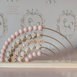 May include: A wooden rainbow abacus with pink beads and geometric shapes. The name "HAZEL HENLE" is spelled out with wooden letter beads. The abacus has a light wood base and gold-colored metal arches.