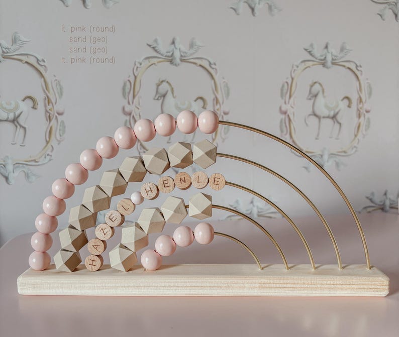 May include: A wooden rainbow abacus with pink beads and geometric shapes. The name "HAZEL HENLE" is spelled out with wooden letter beads. The abacus has a light wood base and gold-colored metal arches.