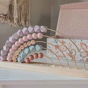 May include: A wooden abacus with a rainbow design. The abacus features rows of lavender, pink, dusty blue, and blush-colored beads. The name "ELEANORA" is spelled out in wooden letters. The base is made of light-colored wood.