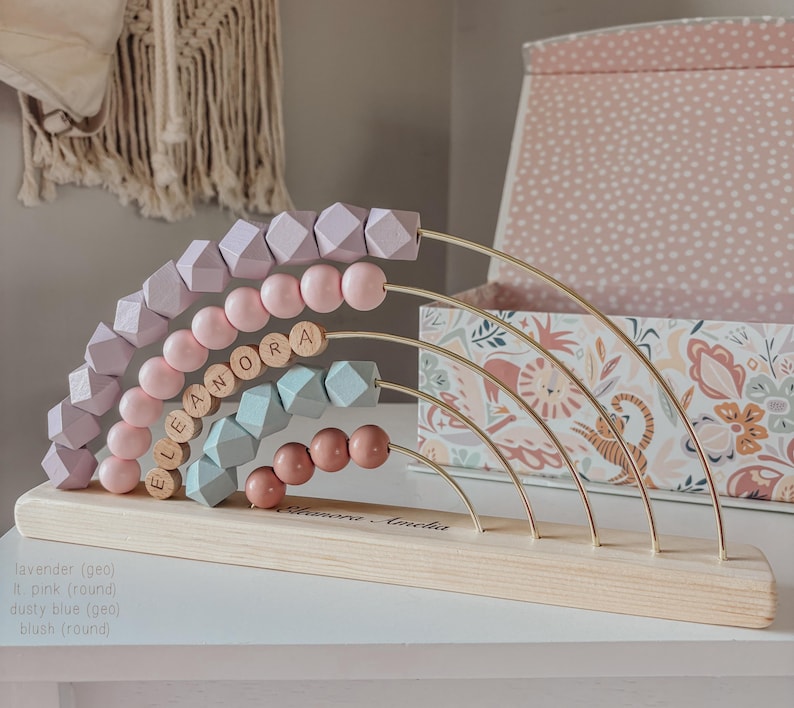 May include: A wooden abacus with a rainbow design. The abacus features rows of lavender, pink, dusty blue, and blush-colored beads. The name "ELEANORA" is spelled out in wooden letters. The base is made of light-colored wood.