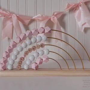 May include: A wooden abacus in a rainbow design with dusty pink and white beads. The abacus has gold-colored arches and the name "Y MARIE E" spelled out with wooden letter beads. The base is made of light-colored wood.