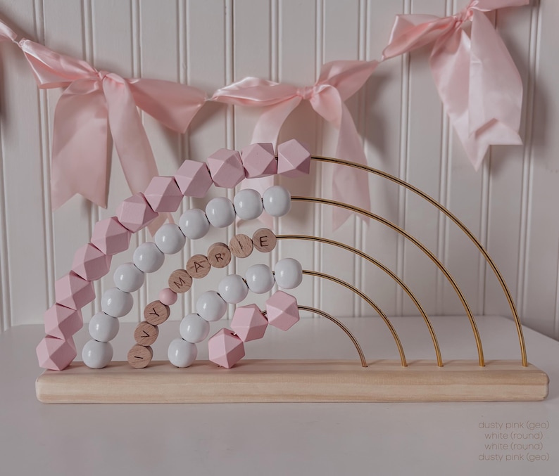 May include: A wooden abacus in a rainbow design with dusty pink and white beads. The abacus has gold-colored arches and the name "Y MARIE E" spelled out with wooden letter beads. The base is made of light-colored wood.
