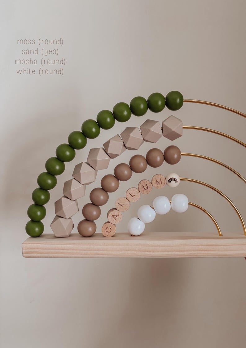 May include: Wooden rainbow-shaped abacus with moss green, sand, mocha, and white beads. The abacus has gold-colored metal arches and a wooden base. The name "CALLUM" is spelled out with wooden letter beads.
