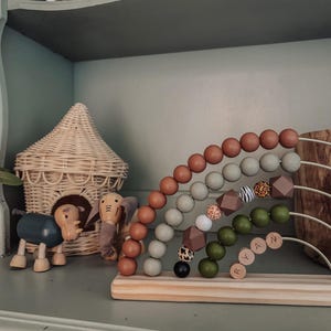 May include: Wooden rainbow abacus with various colored beads, including brown, green, and gray. The abacus has a wooden base with the name "RYAN" spelled out. Also, a wooden elephant and rhino toy, and a woven basket house.