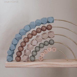 May include: Wooden rainbow-shaped abacus with beads in dusty blue, mocha, sand, and sage colors. The abacus has gold-colored metal arches and a wooden base. The name "MILES" is spelled out with wooden beads.