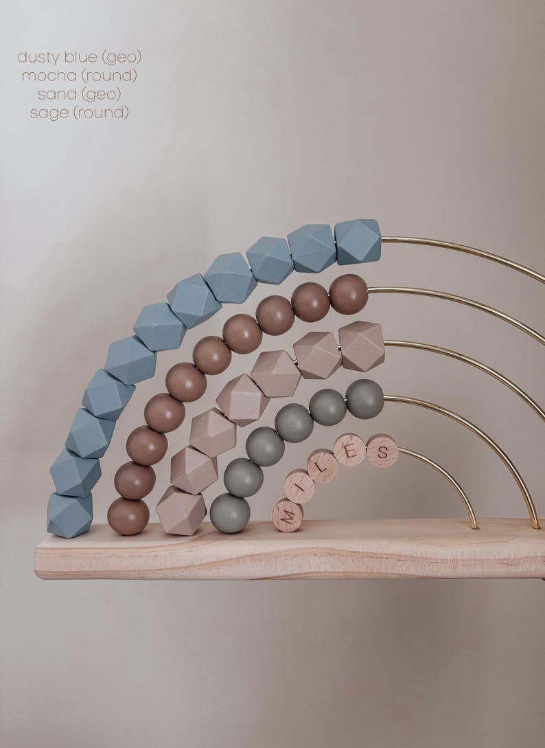 May include: Wooden rainbow-shaped abacus with beads in dusty blue, mocha, sand, and sage colors. The abacus has gold-colored metal arches and a wooden base. The name "MILES" is spelled out with wooden beads.