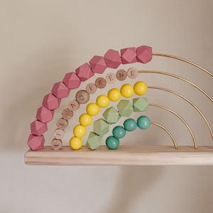 May include: A wooden rainbow-shaped toy with pink, yellow, green, and teal beads. The rainbow has gold metal arches and sits on a wooden shelf. Some beads have names on them.