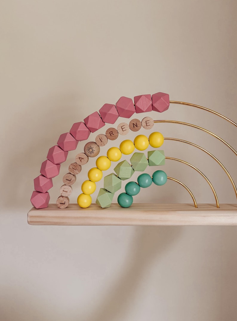 May include: A wooden rainbow-shaped toy with pink, yellow, green, and teal beads. The rainbow has gold metal arches and sits on a wooden shelf. Some beads have names on them.