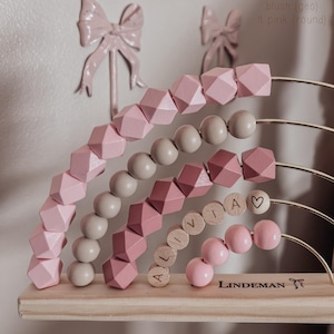 May include: A wooden abacus with pink and sand-colored beads, arranged in a rainbow shape. The name "ALIVIA" is spelled out with wooden letter beads. The base is made of light-colored wood, with the brand name "LINDEMAN" printed on it.