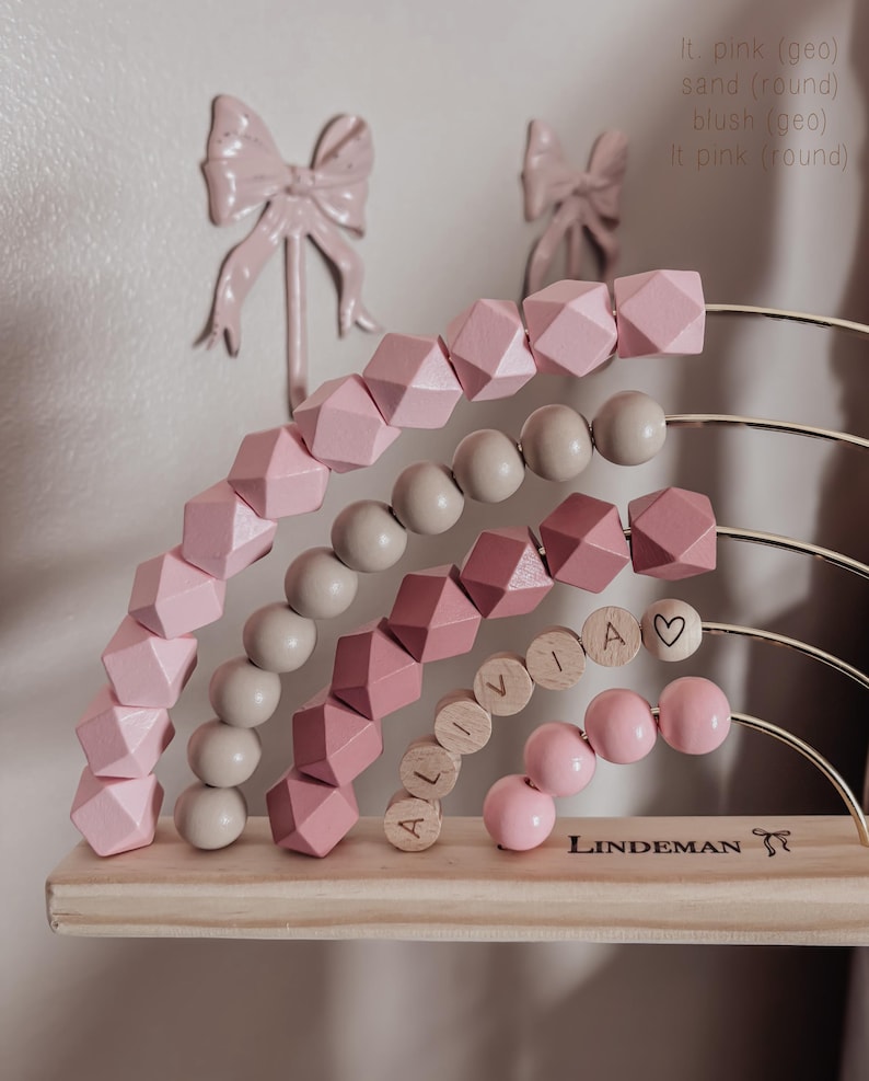 May include: A wooden abacus with pink and sand-colored beads, arranged in a rainbow shape. The name "ALIVIA" is spelled out with wooden letter beads. The base is made of light-colored wood, with the brand name "LINDEMAN" printed on it.