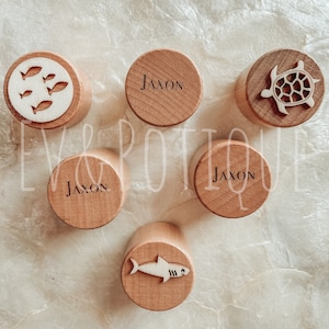 May include: Wooden bottle stoppers with various designs. One stopper features the name "Jaxon" in a simple font. Other stoppers have laser-cut designs of fish, a turtle, and a shark. The stoppers are light brown and cylindrical.