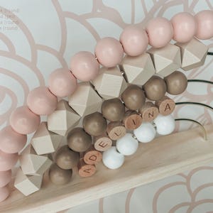 May include: Wooden abacus with light pink, sand, mocha, and white beads. The abacus has round and geometric shapes, with the name "ADELINA" spelled out in wooden letters. The frame is light wood with gold-colored metal arches.