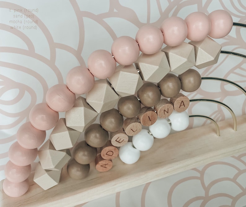 May include: Wooden abacus with light pink, sand, mocha, and white beads. The abacus has round and geometric shapes, with the name "ADELINA" spelled out in wooden letters. The frame is light wood with gold-colored metal arches.