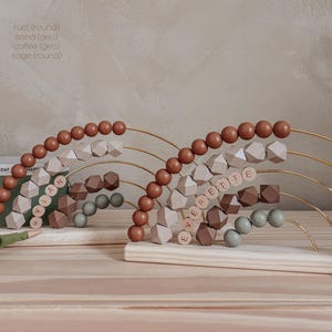May include: Wooden abacus with rust, sand, coffee, and sage colored beads. The abacus has a rainbow design with the names "LANAH" and "EVERETIE" spelled out. A wooden giraffe and crocodile are also present.