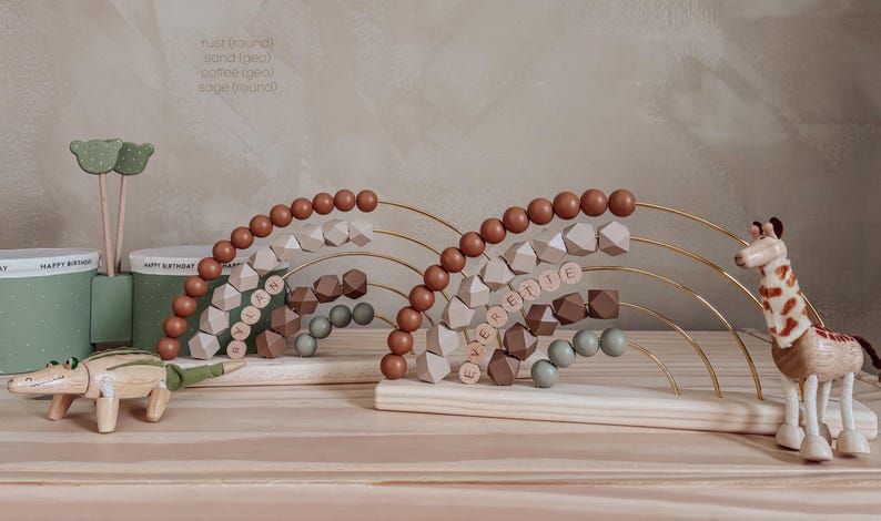 May include: Wooden abacus with rust, sand, coffee, and sage colored beads. The abacus has a rainbow design with the names "LANAH" and "EVERETIE" spelled out. A wooden giraffe and crocodile are also present.