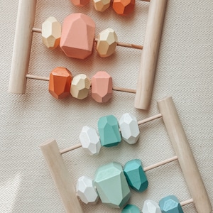 May include: Wooden abacuses with colorful, geometric beads. One abacus features peach, orange, and yellow beads. The other has white, teal, and blue beads. The frames are made of natural wood.