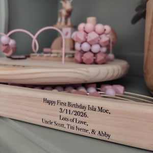 May include: Wooden toy set with pink and light brown elements, including a bead maze and geometric shapes. The text on the wooden base reads "Happy First Birthday Isla Moe, 3/11/2026. Lots of Love, Uncle Scott, Titi Ivette, & Abby."