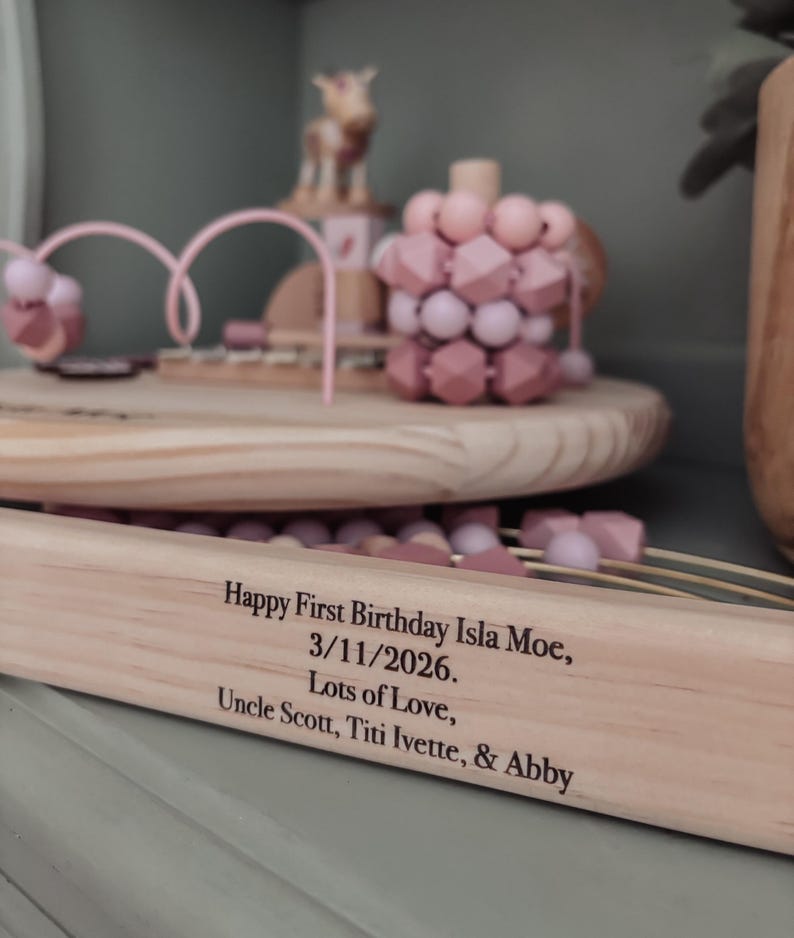 May include: Wooden toy set with pink and light brown elements, including a bead maze and geometric shapes. The text on the wooden base reads "Happy First Birthday Isla Moe, 3/11/2026. Lots of Love, Uncle Scott, Titi Ivette, & Abby."