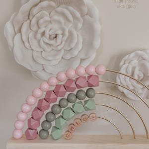 May include: A wooden rainbow-shaped abacus with pink, mauve, sage green, and gray beads. The name "CHLOE" is spelled out with wooden letter blocks. The background features a large white floral wall decoration.