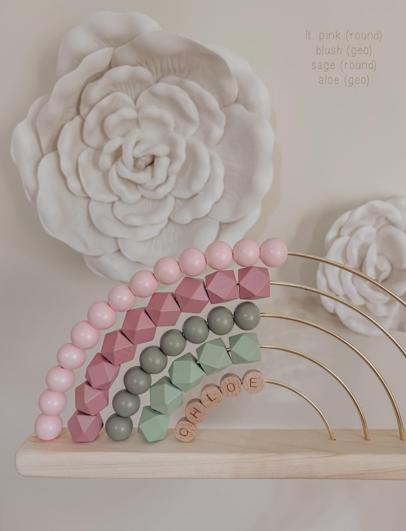May include: A wooden rainbow-shaped abacus with pink, mauve, sage green, and gray beads. The name "CHLOE" is spelled out with wooden letter blocks. The background features a large white floral wall decoration.