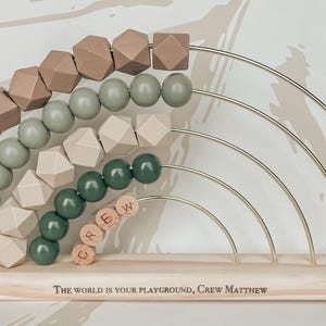 May include: Wooden rainbow abacus with geometric and round beads in shades of brown, green, and beige. The abacus has a wooden base with the text "The world is your playground, Crew Matthew". The beads are strung on metal arches.