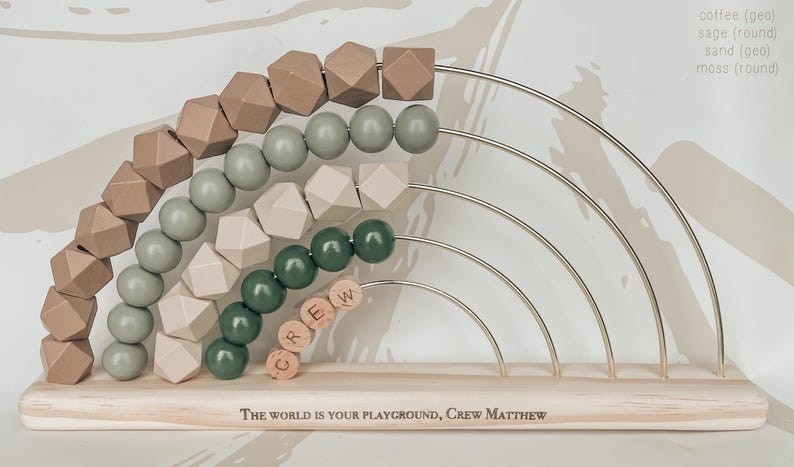 May include: Wooden rainbow abacus with geometric and round beads in shades of brown, green, and beige. The abacus has a wooden base with the text "The world is your playground, Crew Matthew". The beads are strung on metal arches.