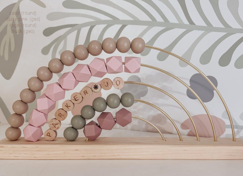 May include: Wooden abacus toy with a rainbow design. The abacus features beads in shades of sand, dusty pink, sage, and blush. The name "PARKER JO" is spelled out with wooden letter beads. The toy is set on a wooden base.