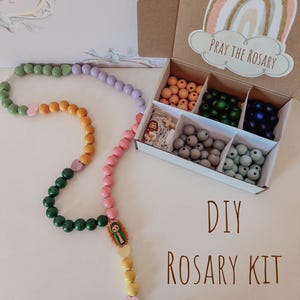 May include: A DIY rosary kit featuring a cardboard box with compartments filled with assorted colored wooden beads, a small Jesus figure, and a wooden cross. A partially assembled rosary with green, yellow, pink, and lavender beads is displayed.
