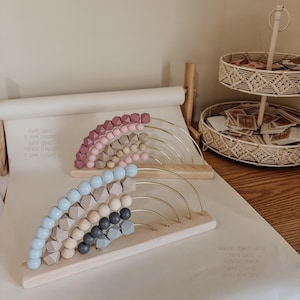 May include: Two wooden abacuses with rainbow-colored beads. One has pink and tan beads, the other has blue, gray, and natural-colored beads. The beads are arranged on gold-colored arches attached to a wooden base.