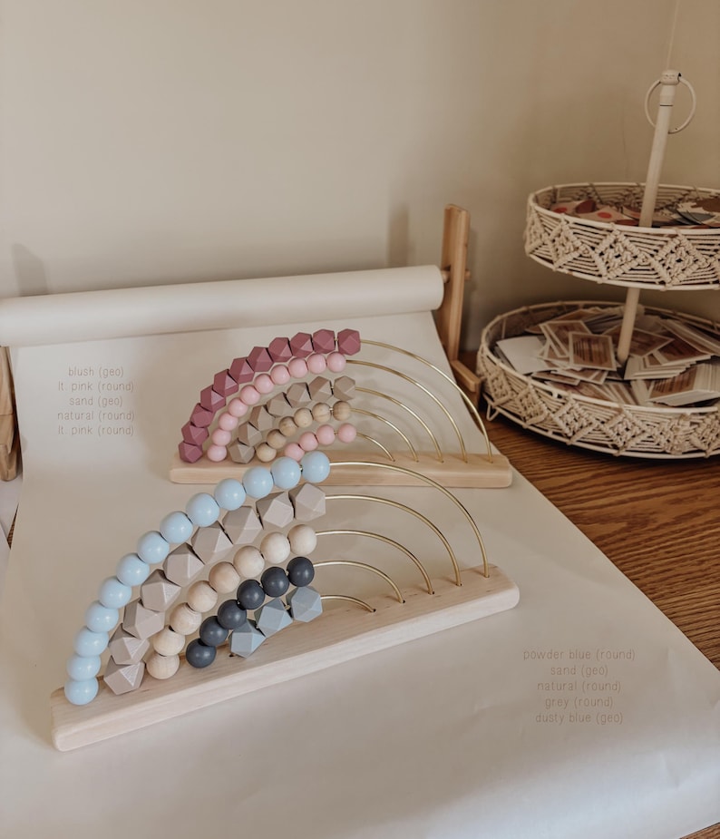 May include: Two wooden abacuses with rainbow-colored beads. One has pink and tan beads, the other has blue, gray, and natural-colored beads. The beads are arranged on gold-colored arches attached to a wooden base.