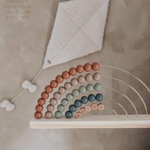 May include: A wooden rainbow abacus with rust, mocha, sage, and grey beads. The abacus sits on a wooden shelf, with the name "ROCCO" spelled out in wooden letters. A white kite-shaped wall hanging is above the abacus.