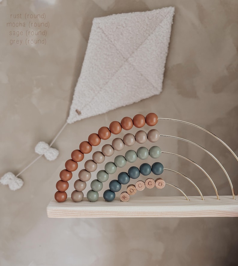 May include: A wooden rainbow abacus with rust, mocha, sage, and grey beads. The abacus sits on a wooden shelf, with the name "ROCCO" spelled out in wooden letters. A white kite-shaped wall hanging is above the abacus.