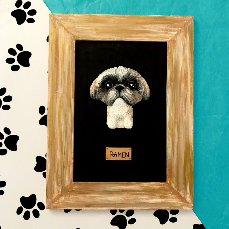 May include: A framed portrait of a Shih Tzu dog with the word "Ramen" on a small plaque below the dog. The frame is a light brown wood with a black background.