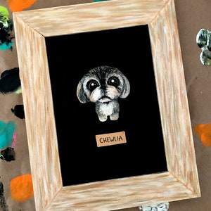 May include: A framed painting of a small, gray dog with big eyes. The dog is painted in a cartoon style and is looking directly at the viewer. The frame is a light brown wood with a white wash. The name "CHEWLIA" is written on a small, brown tag below the dog.