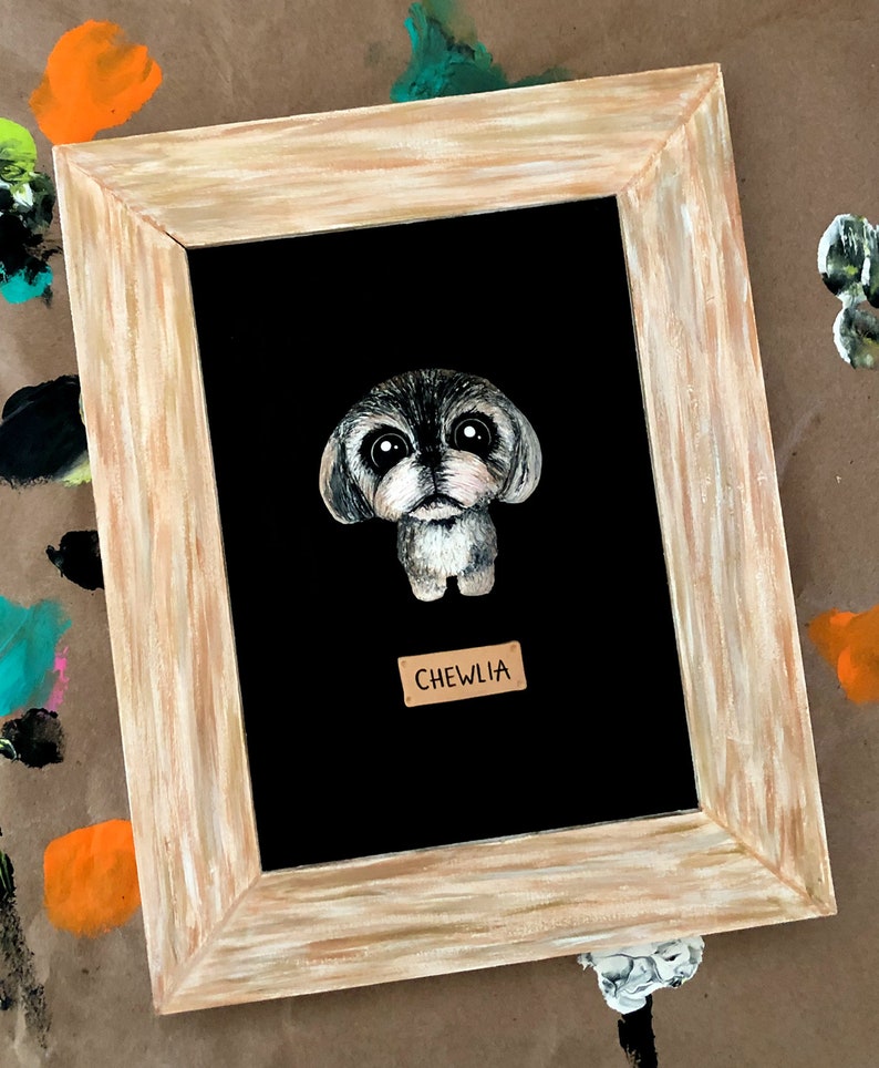 May include: A framed painting of a small, gray dog with big eyes. The dog is painted in a cartoon style and is looking directly at the viewer. The frame is a light brown wood with a white wash. The name "CHEWLIA" is written on a small, brown tag below the dog.