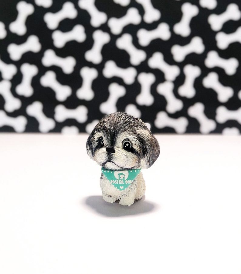 May include: A small, gray and white, hand-painted figurine of a Shih Tzu dog wearing a teal blue bandana with white polka dots and the text "Foster Dogs".