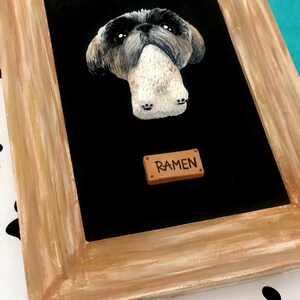 May include: A framed portrait of a small, white and gray dog with black eyes. The dog is in a sitting position with its paws visible. The frame is made of wood and has a brown stain. A small brown plaque with the name "RAMEN" is attached to the bottom of the frame.